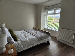 A bedroom with a bed, a window with blinds, a radiator, and a bedside table with a lamp and a vase. - 8 Arvonia Terrace (Criccieth)