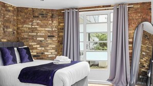 A bedroom with a brick wall, a bed with white bedding and purple accents, a window with curtains, and a patterned rug. - 5 Bed/2 Bath Private House with Balcony - London (London)