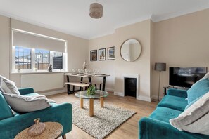 Ein Wohnzimmer mit petrolfarbenen Sofas, einem Glastisch, einem Kamin, einem Fernseher und einem Essbereich mit Tisch und Stühlen.