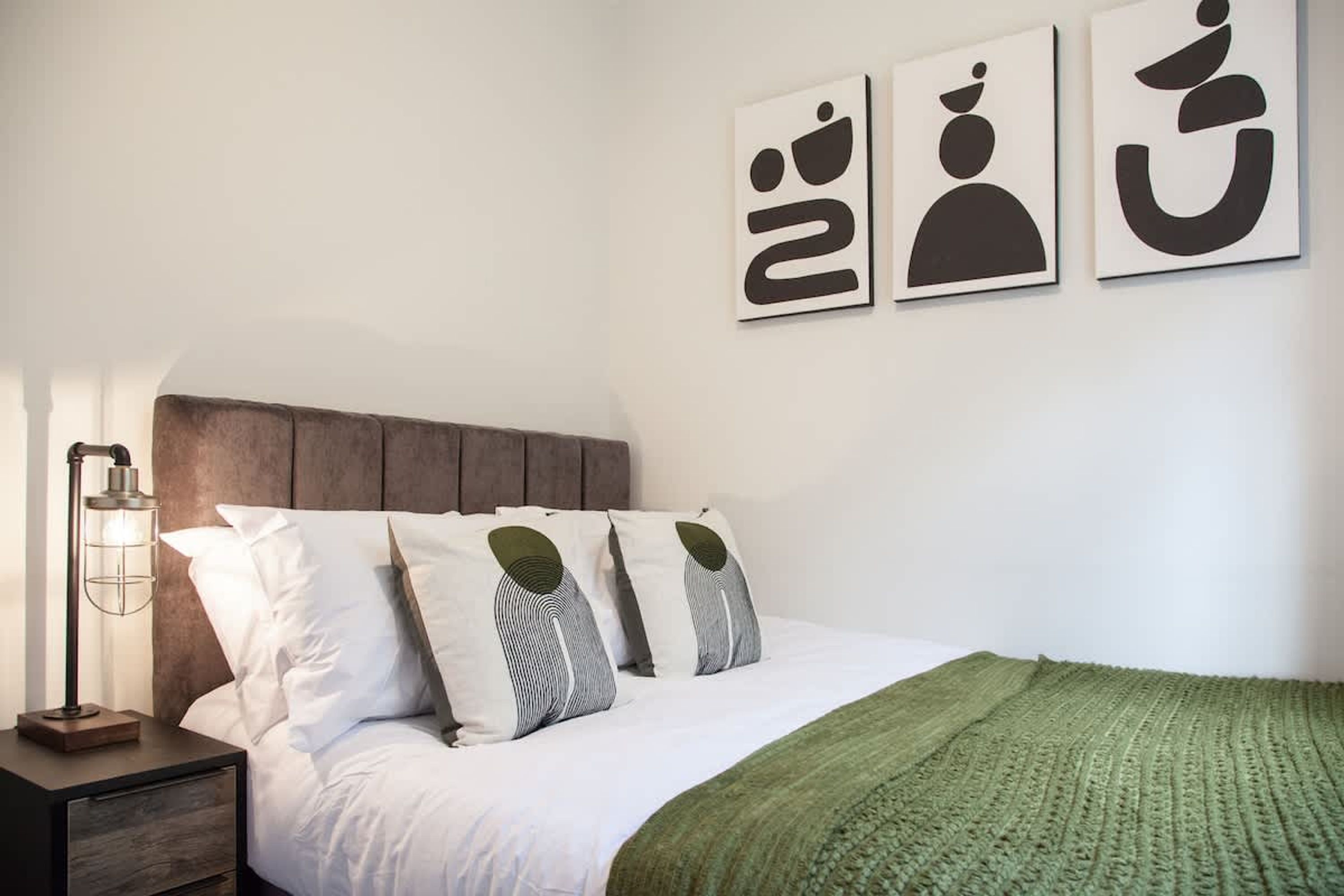 Ein ordentlich gemachtes Bett mit weißer Bettwäsche und einer grünen Decke, eine Nachttischlampe sowie drei abstrakte Wandbilder.