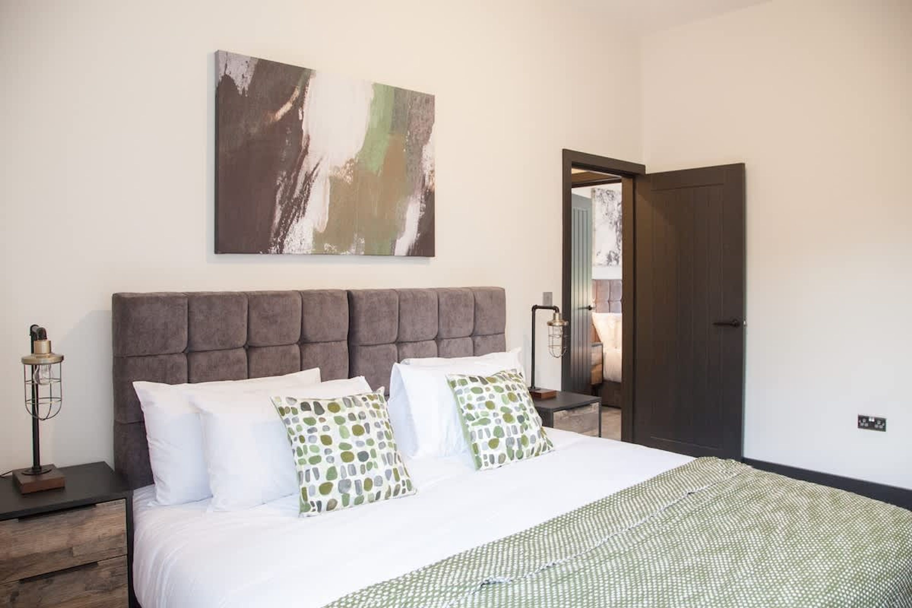 Un dormitorio con una cama grande, cabecera, mesita de noche con lámpara, un cuadro en la pared y una puerta que da a otra habitación.