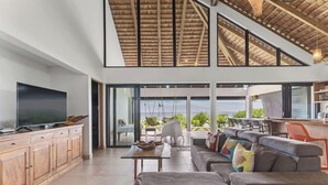 A spacious living area with a large TV, wooden cabinets, a sofa, and a dining area with a view. - Coastal Bliss 1BR Bungalow by the Sea in Moorea (Moorea-Maiao)