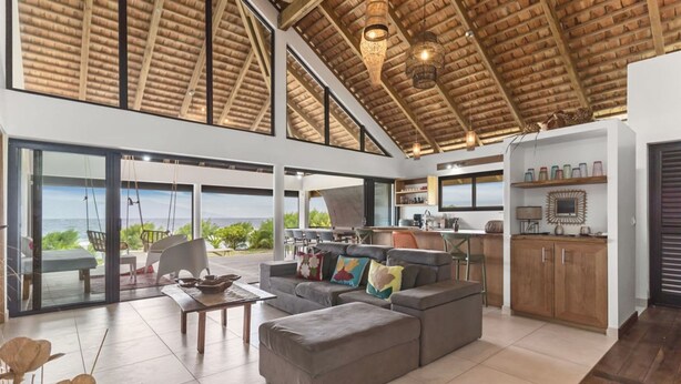 A spacious living area with a large sofa, a dining table, and a kitchen area with a view. - Coastal Bliss 1BR Bungalow by the Sea in Moorea (Moorea-Maiao)