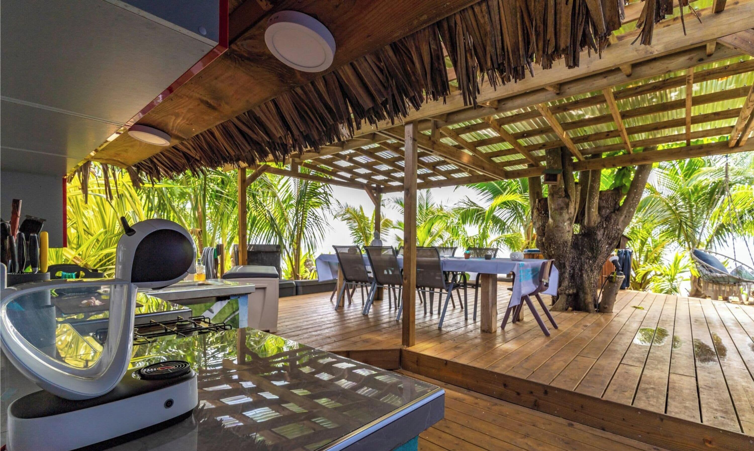 An outdoor dining area with a grill, tables, chairs, and a thatched roof structure.