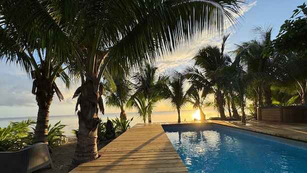 A wooden deck leading to a swimming pool with palm trees and a sunset view.