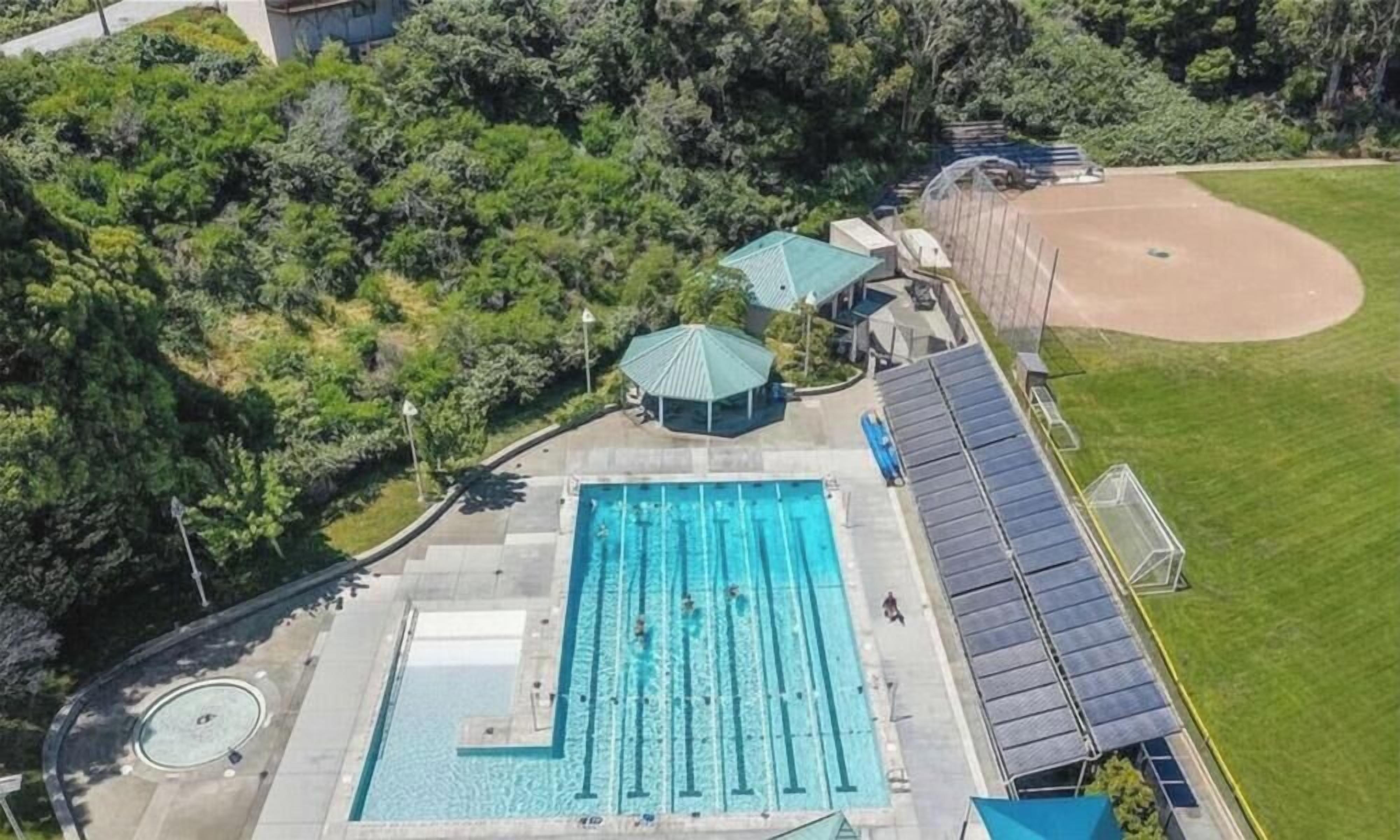 Ovenfra ses en swimmingpool med tilhørende dæk, en baseballbane og et overdækket siddeområde.