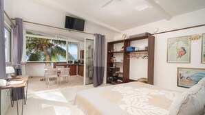 A bedroom with a bed, a desk, a chair, a TV, a shelf, and a view of palm trees. - Seaside Serenity 2BR Oasis on Enchanting Moorea (Moorea-Maiao)