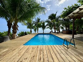 Una terraza de madera con piscina, palmeras y vista al océano.