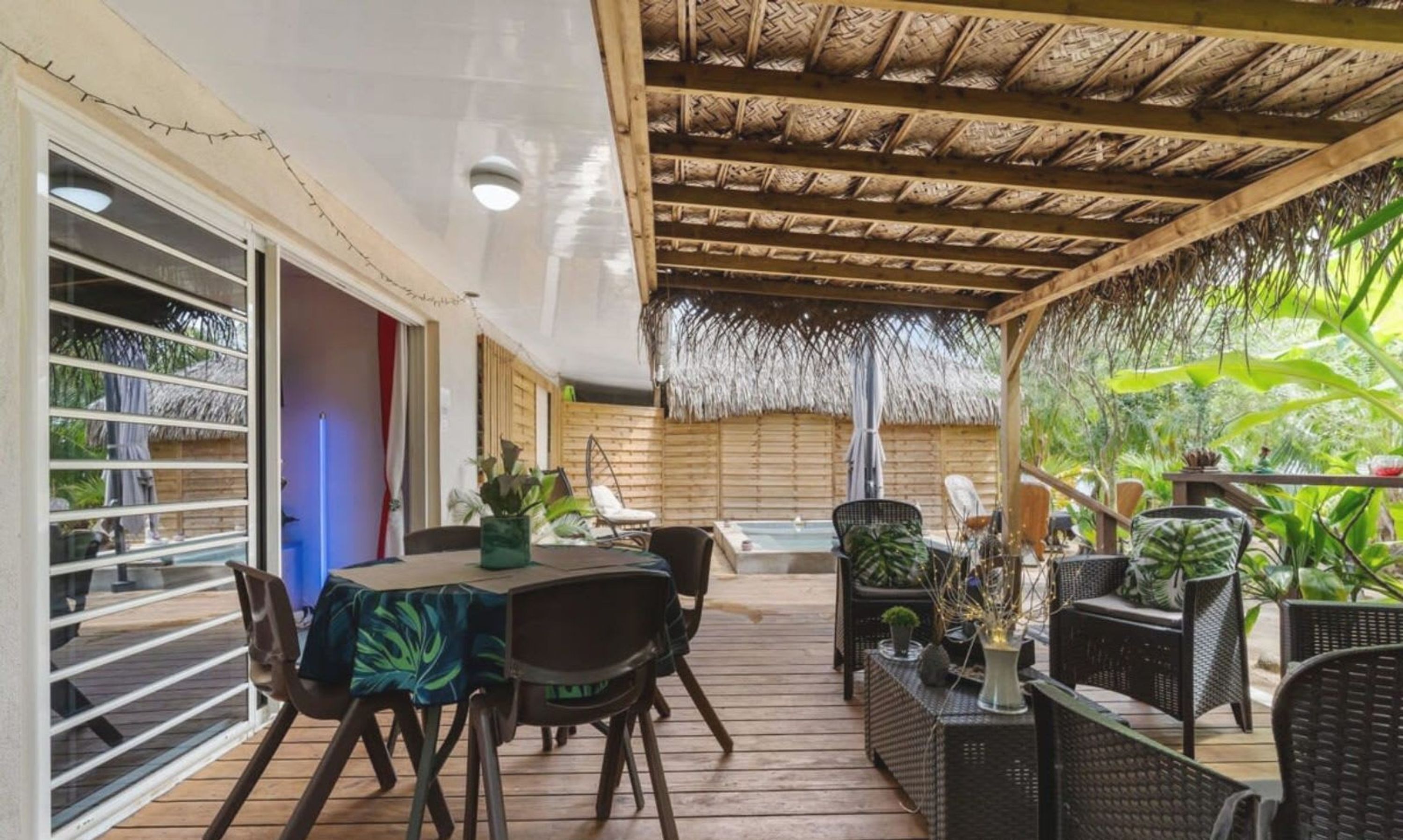 Une terrasse extérieure couverte, dotée d’une table et de chaises en bois, entourée de plantes en pot et surmontée d’un toit de chaume.