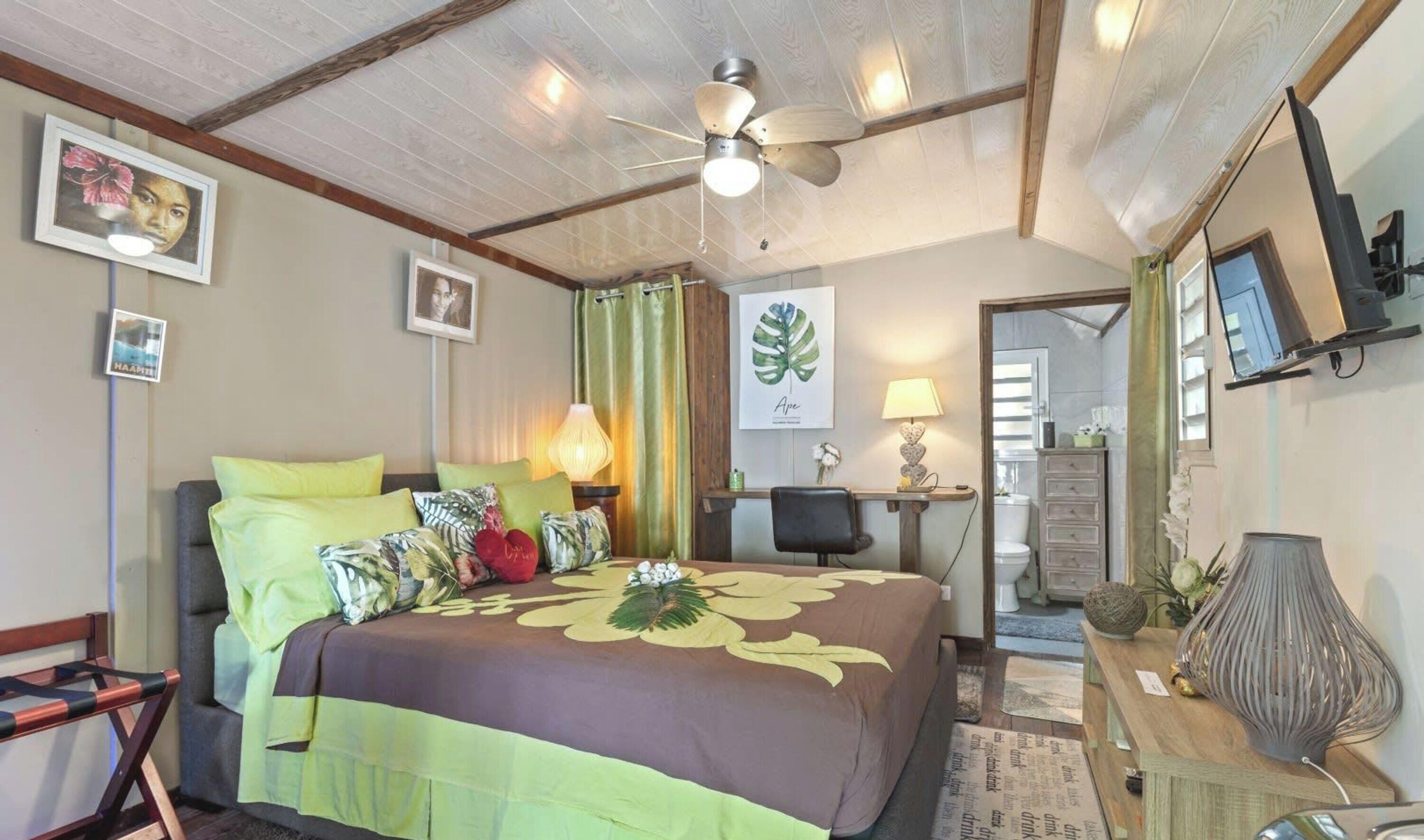 Een slaapkamer met een bed, een bureau, een stoel, een plafondventilator en een zichtbare badkamer door een geopende deur.