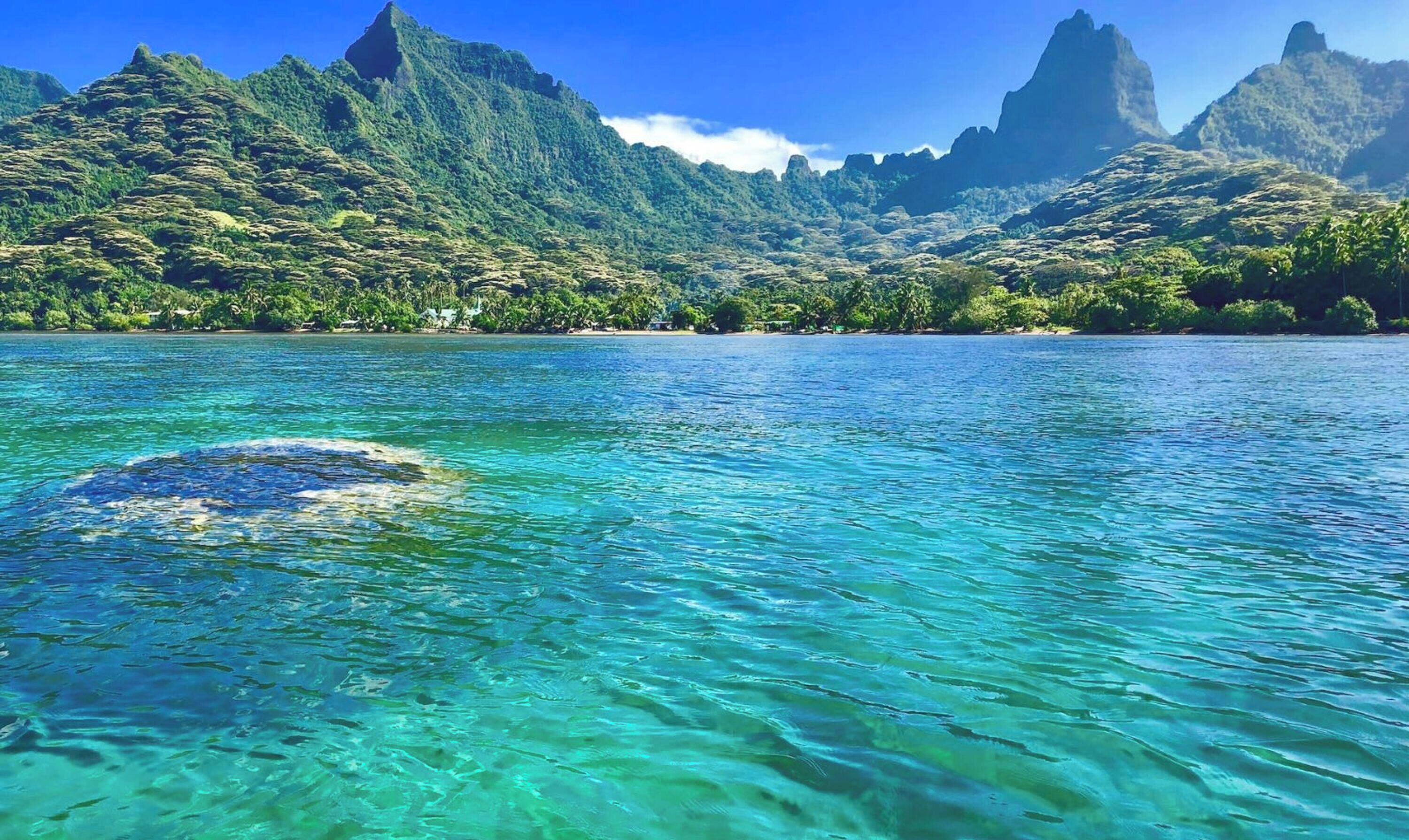 Mar tropical cristalina con una isla rocosa en primer plano y montañas verdes exuberantes al fondo.