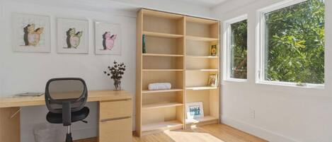 Una stanza con una scrivania in legno, una sedia da ufficio nera, una libreria e una finestra vista sul verde.