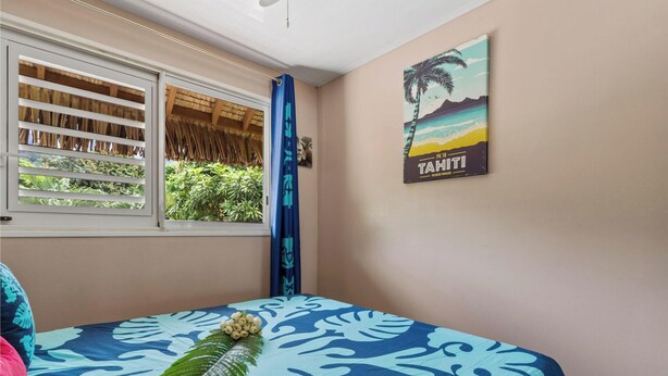 A bedroom with a bed, a window with shutters, a tropical-themed wall poster, and a view of greenery outside. - Beachfront Bungalows With Sunset Views in Haapiti (Moorea-Maiao)