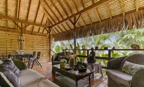 A covered outdoor seating area with wicker furniture, a table with decorative items, and a view of palm trees. - Beachfront Bungalows With Sunset Views in Haapiti (Moorea-Maiao)