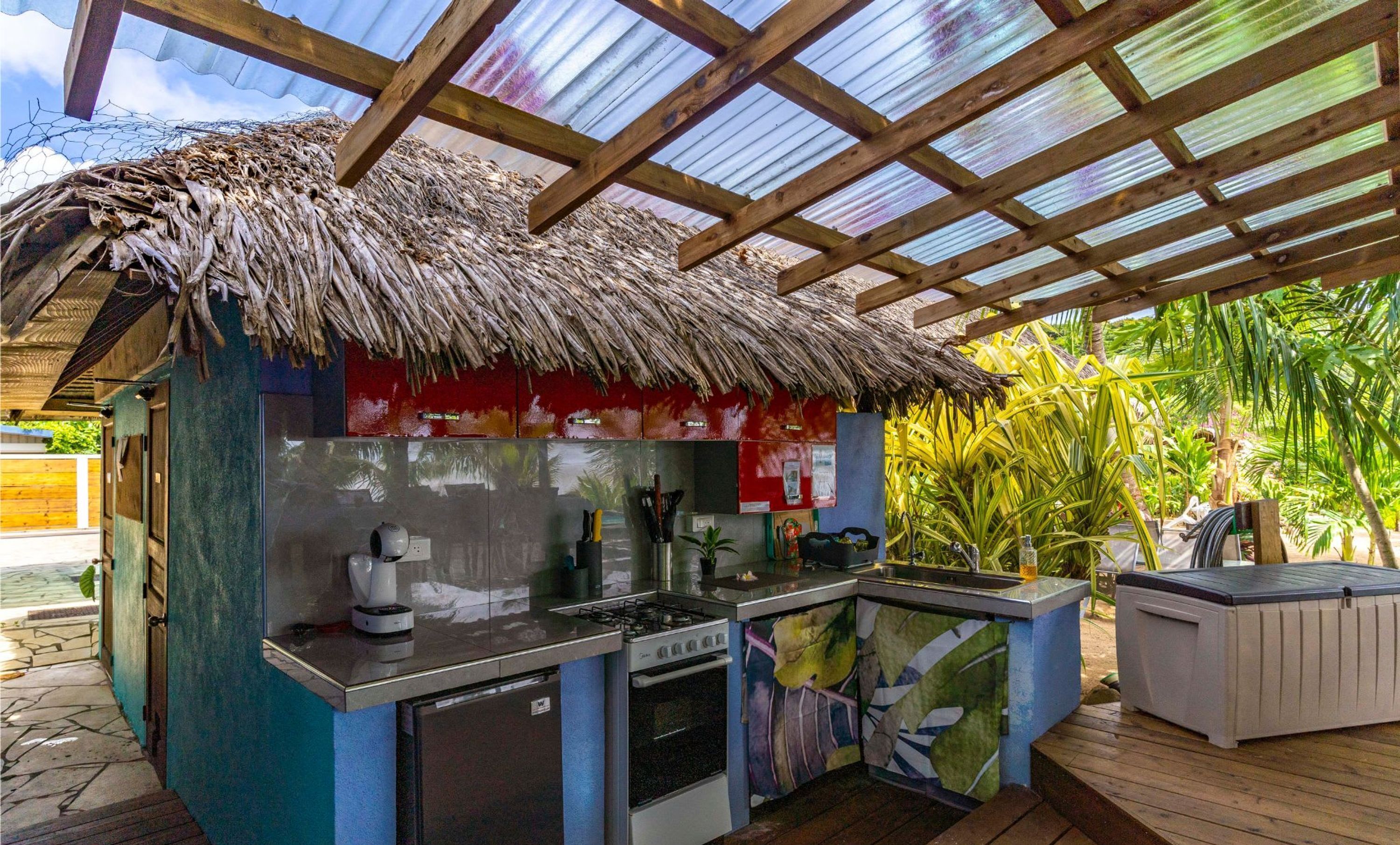 A thatched-roof outdoor kitchen with a gas stove, refrigerator, and a colorful leaf-patterned backsplash.