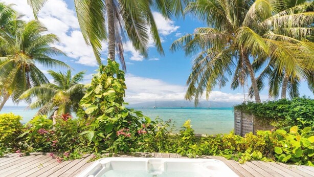 Terraza de madera con un jacuzzi, rodeada de exuberante follaje y palmeras, con vistas al mar azul y transparente.
