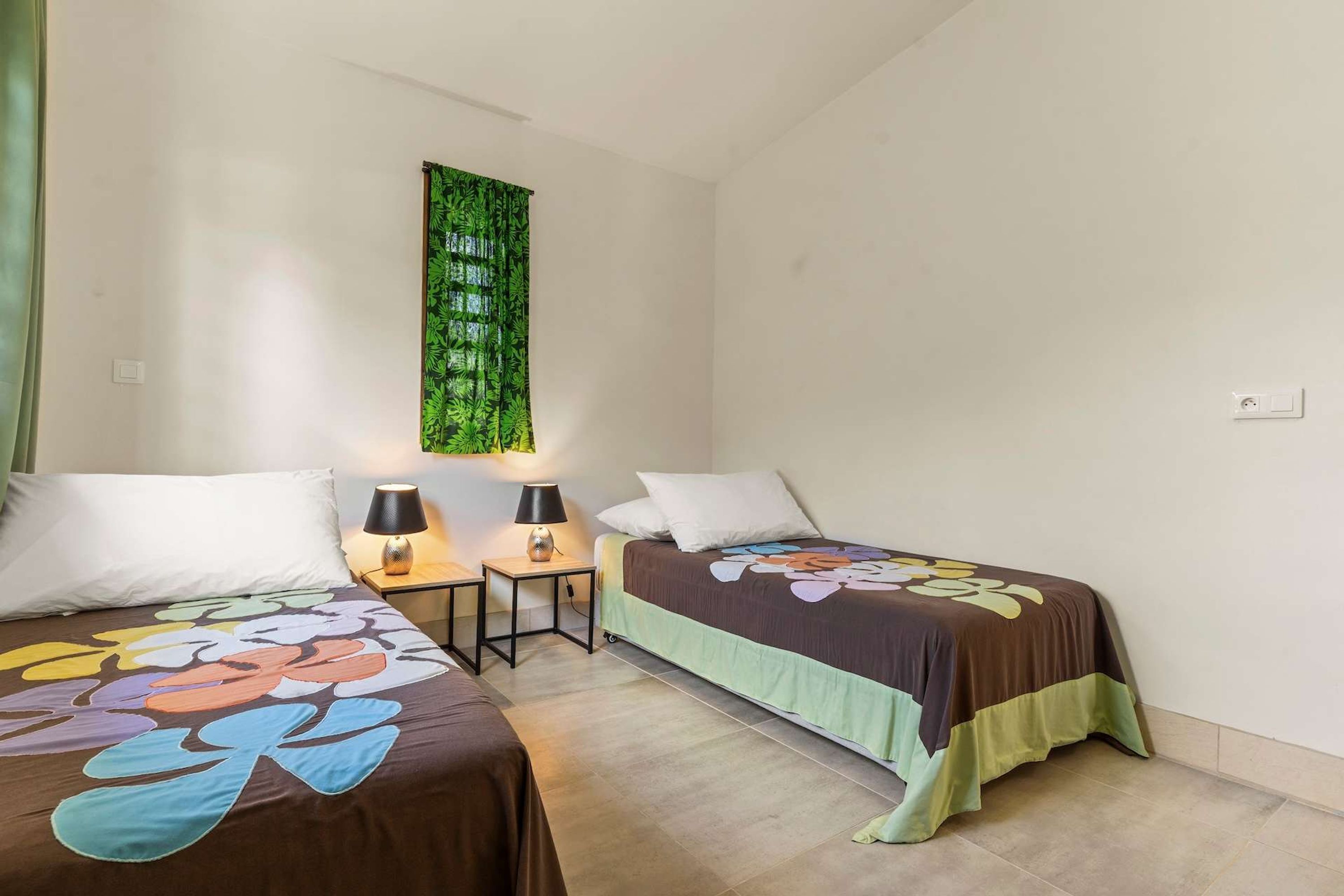 A room with two beds, a nightstand with a lamp, a wall-mounted light, and a green curtain.
