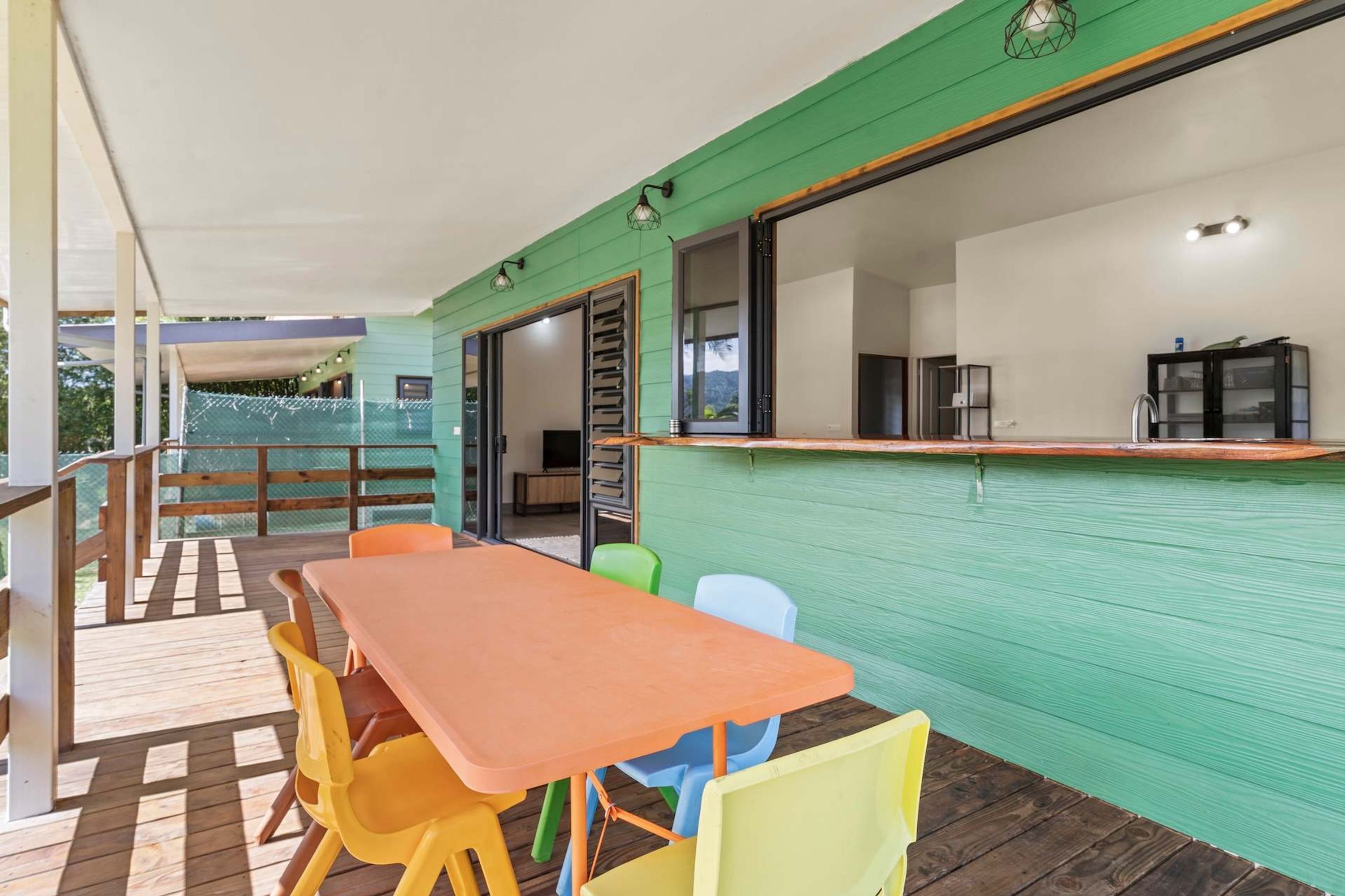 Un espace extérieur abrité, aménagé pour les repas, avec une table en bois et des chaises colorées, un coin cuisine doté d’un comptoir et de placards, et une vue sur un paysage verdoyant à travers une clôture.
