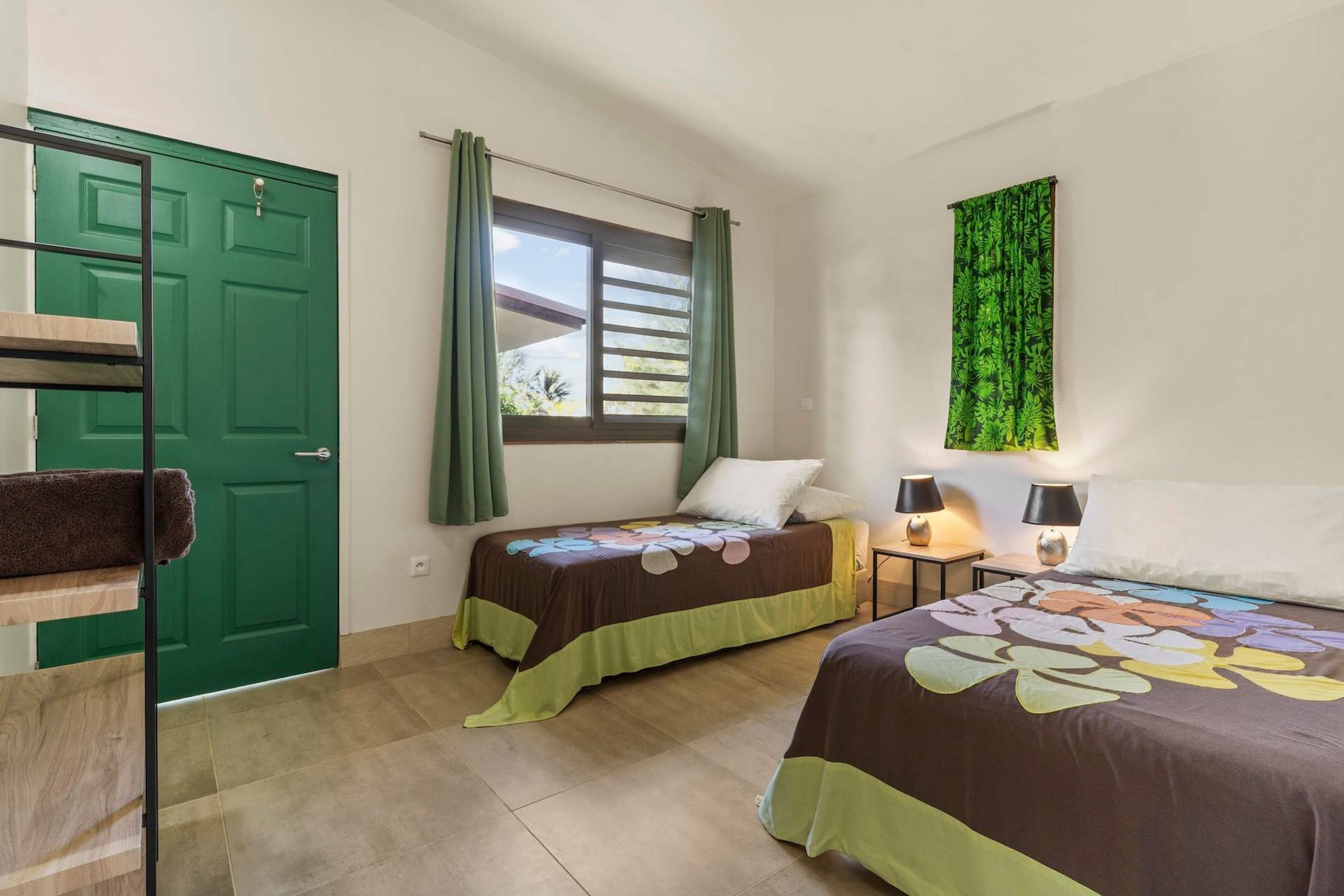 Una habitación con dos camas, una puerta verde, una ventana con cortinas y una planta colgada en la pared.