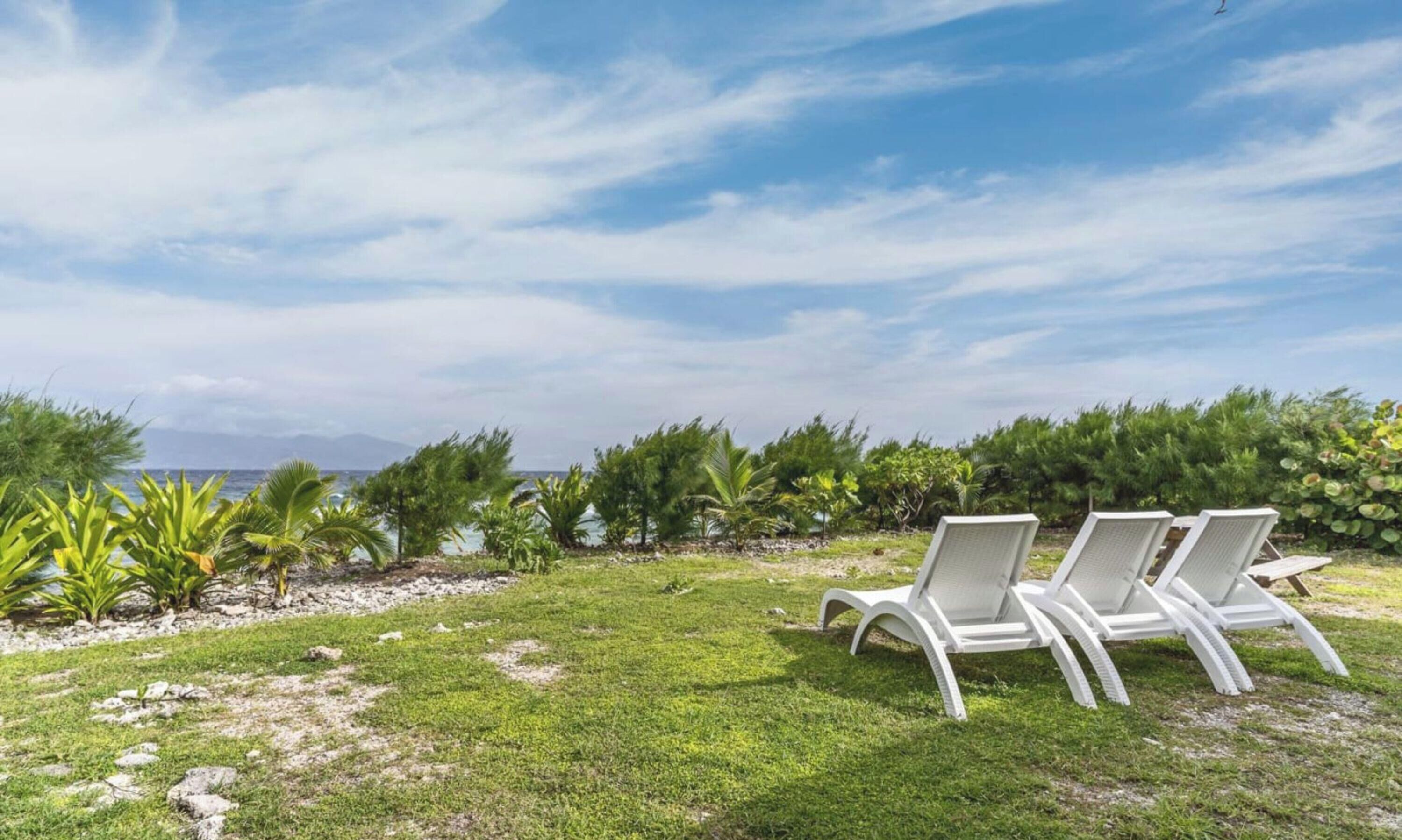 A grassy area with three white lounge chairs facing a body of water and a clear sky.