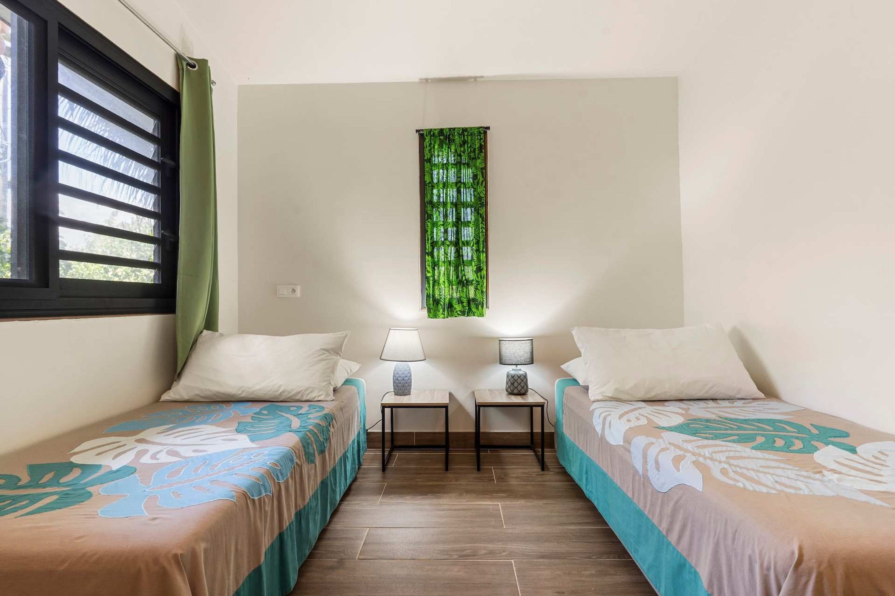A room with two beds, a window with blinds, a green curtain, a wall-mounted green tapestry, and two bedside tables with lamps.