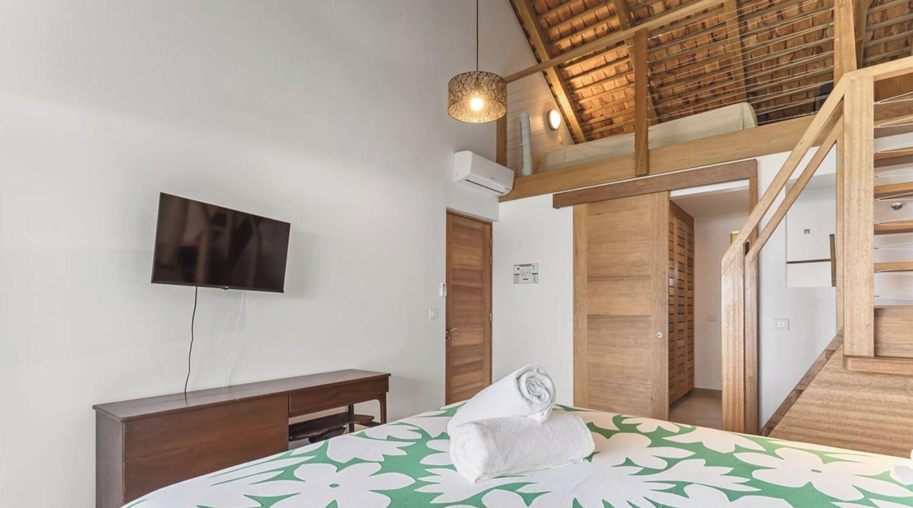 A bedroom with a bed, a TV, a wooden desk, a staircase, and an air conditioning unit.