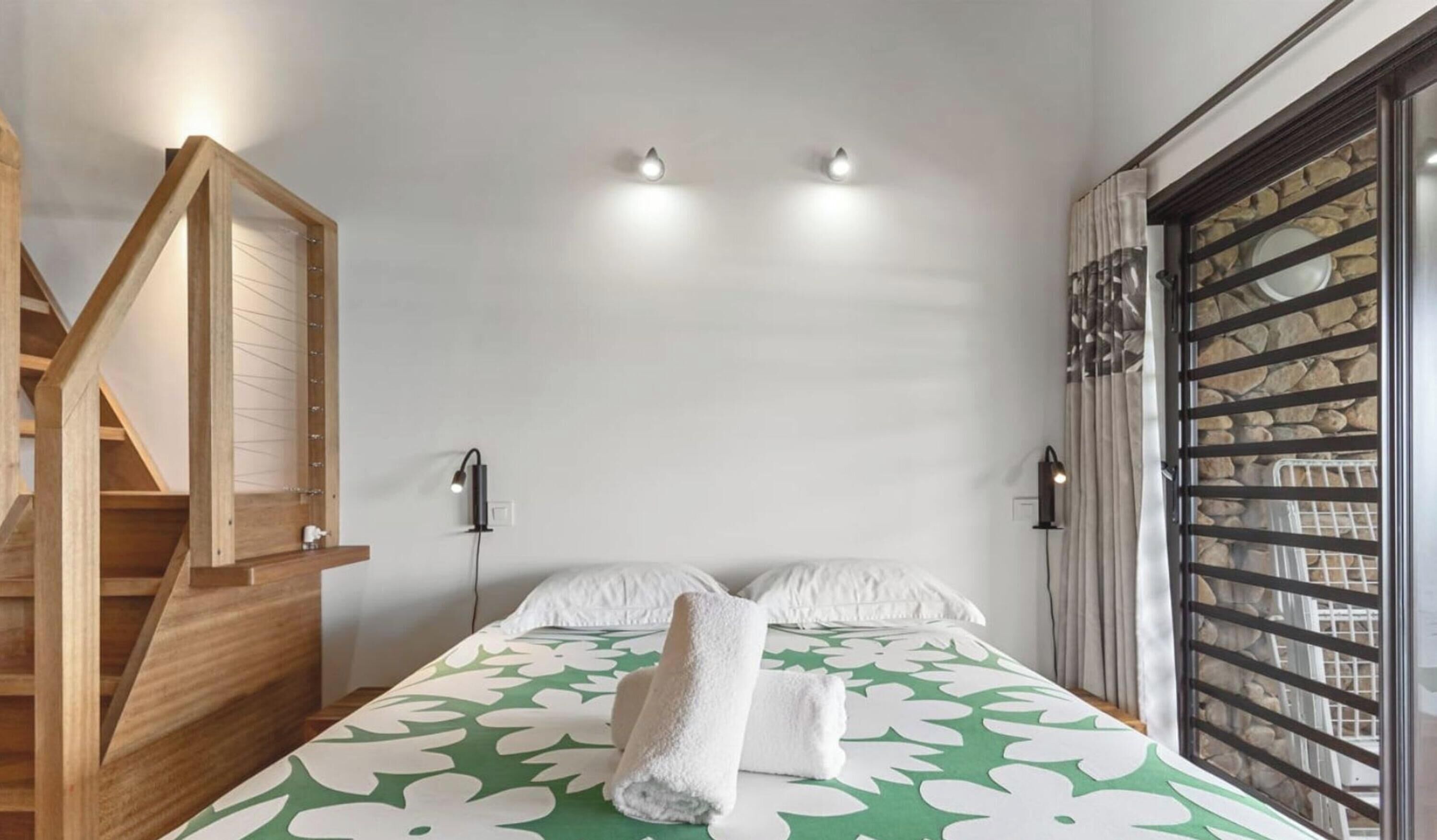 Un dormitorio con cama, dos lámparas de noche, una escalera de madera y una ventana con pared de piedra.