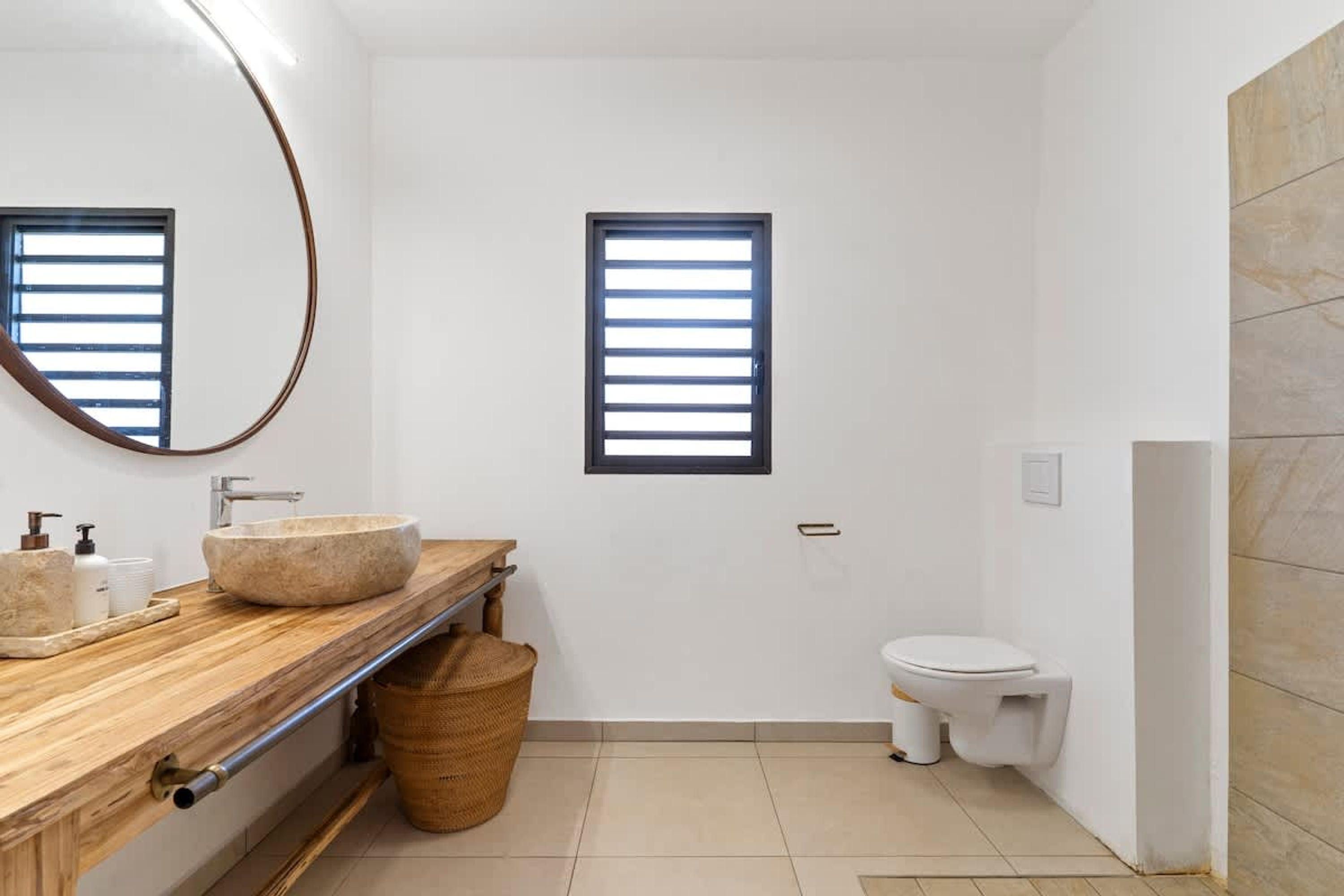 A modern bathroom with a wooden vanity, a round sink, a toilet, and a window with blinds.