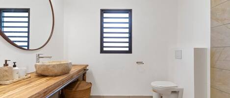 A modern bathroom with a wooden vanity, a round sink, a toilet, and a window with blinds.
