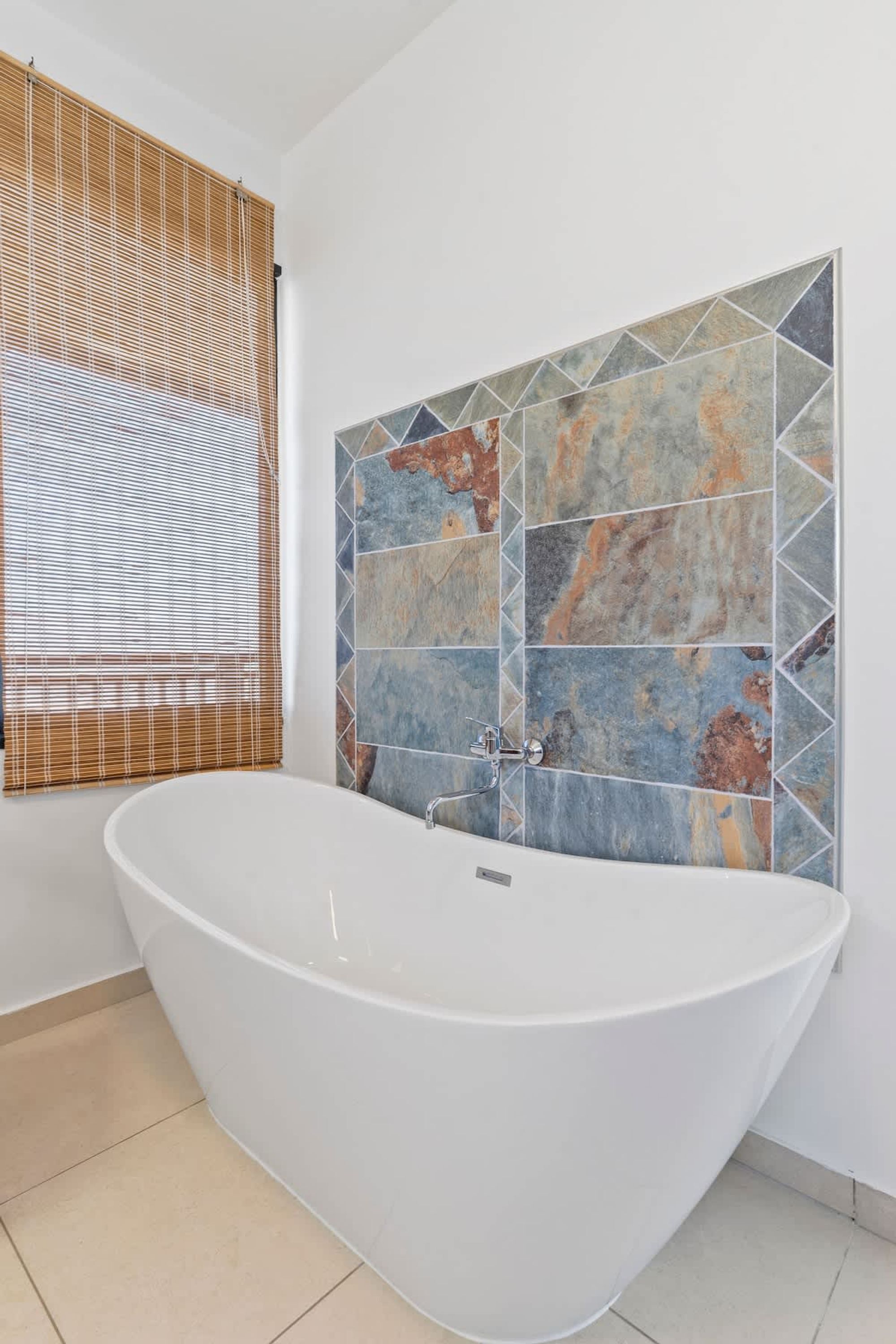 Een badkamer met een vrijstaand bad, een raam met zonwering en een wand met een geometrisch tegelpatroon.