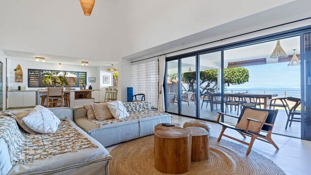 Un salon moderne comprenant un canapé, une table basse et des fauteuils, un coin cuisine et une grande porte-fenêtre coulissante donnant sur un espace repas extérieur avec vue sur la mer.