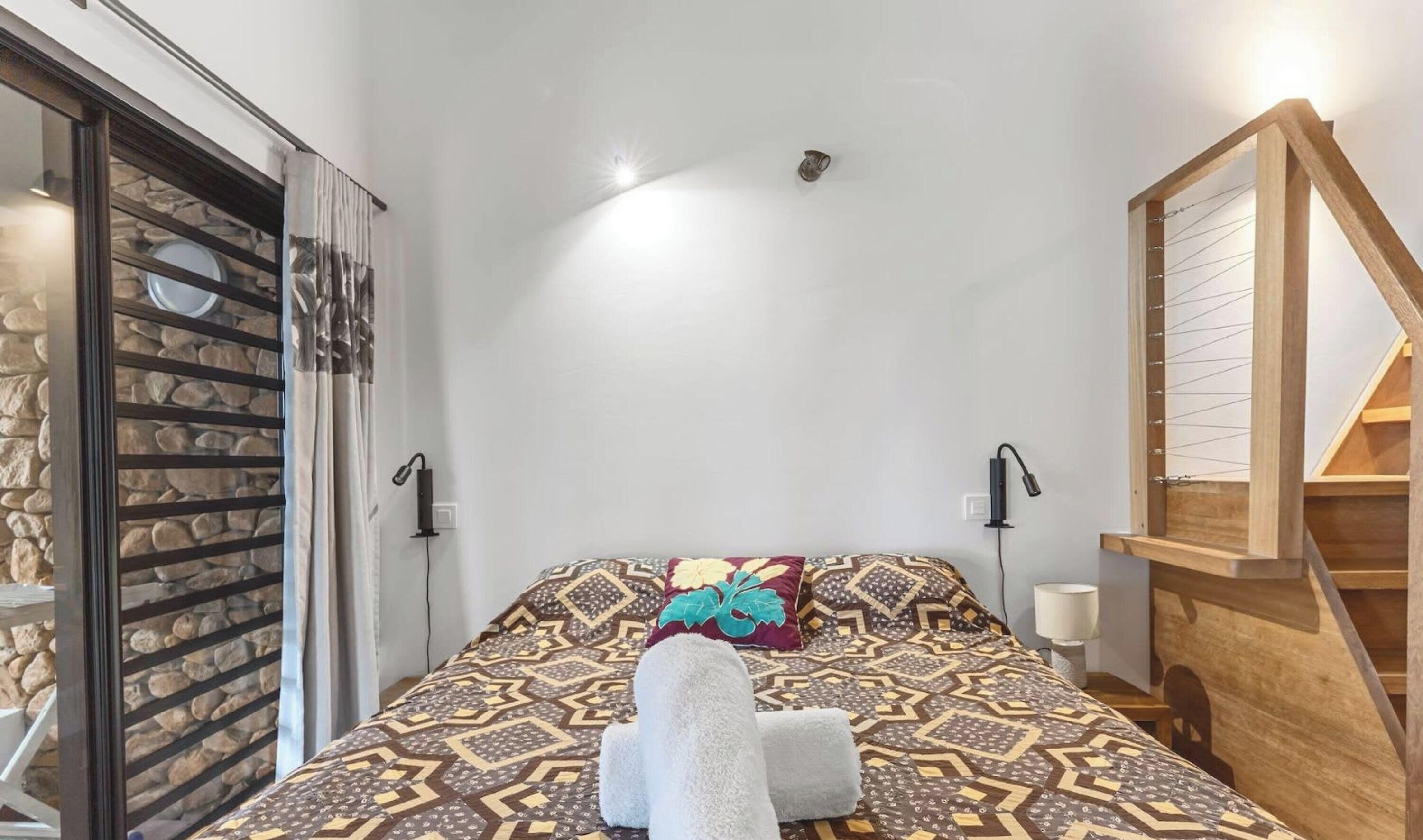 A bedroom with a patterned bedspread, a pair of towels, a wooden staircase, and a stone wall.
