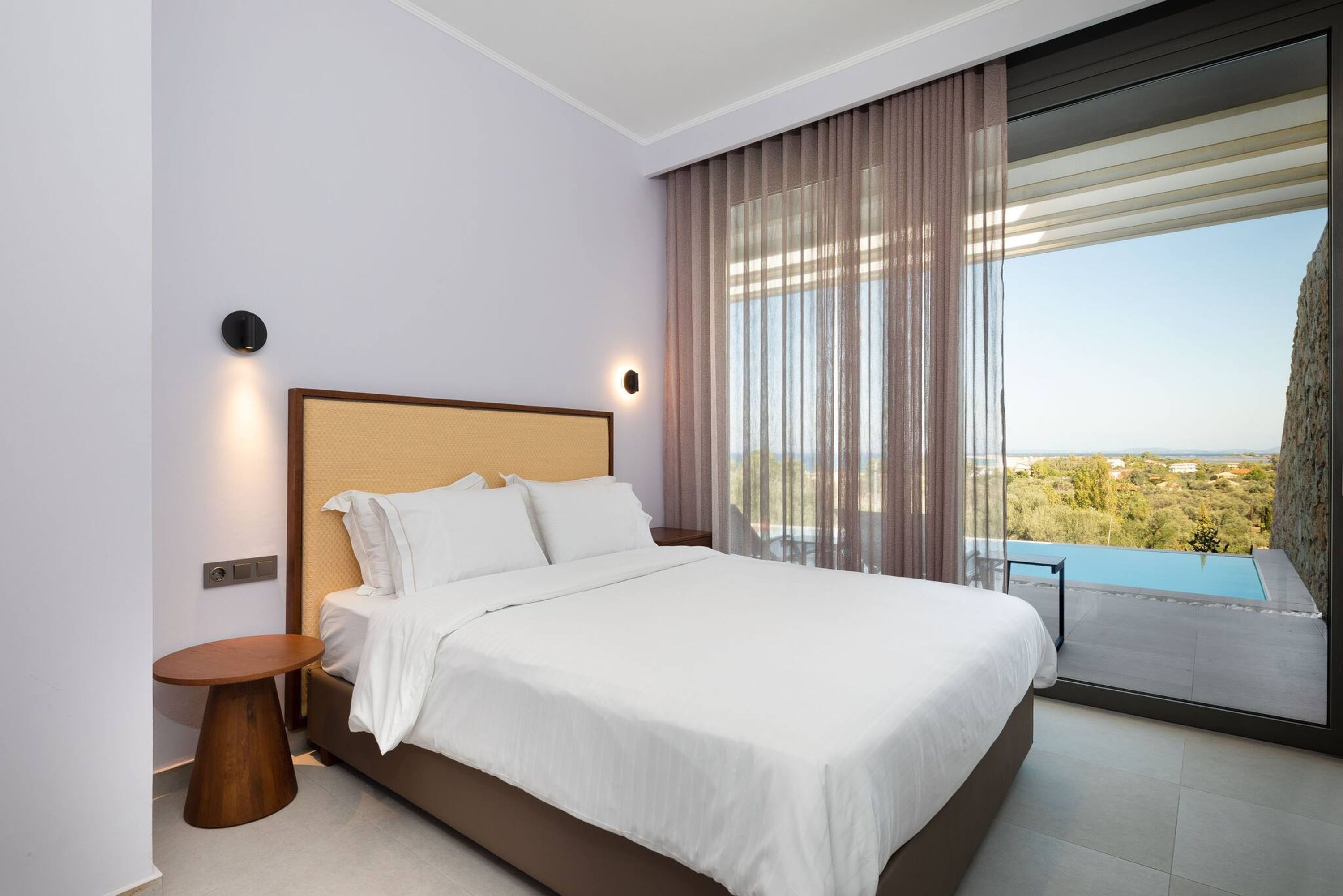 Ein modernes Schlafzimmer mit einem großen Bett, einem kleinen runden Tisch und Blick auf einen Pool und die Landschaft.