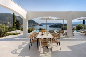 A covered outdoor dining area with a table, chairs, and umbrellas, overlooking a lake and mountains. - Villa Unique in Lefkada (Lefkada)