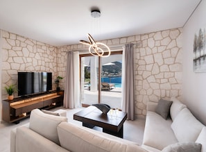A modern living room with a stone wall, a flat-screen TV, a sofa, a coffee table, and a view of a pool and mountains. - Villa Unique in Lefkada (Lefkada)