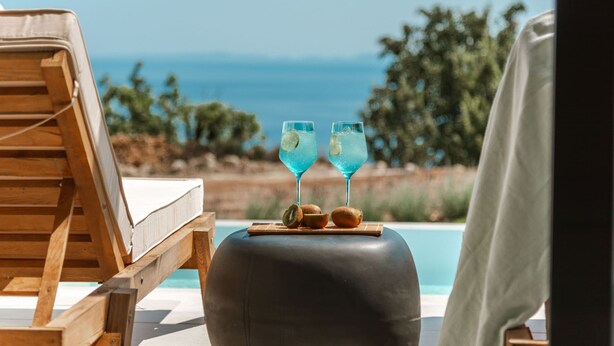 A wooden lounge chair by a pool with two glasses of blue drink and kiwi slices on a round ottoman. - Orizontas 1 Syvota in Igoumenitsa (Igoumenitsa)
