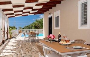 A covered outdoor dining area with a stone floor, white furniture, and a view of a swimming pool and greenery. - Dream 1 BED in Ponti (Lefkada)