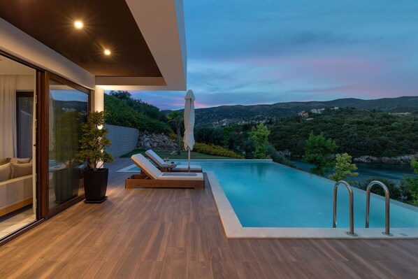 A modern outdoor pool area with a view of a river and hills, wooden deck, and lounge chairs.
