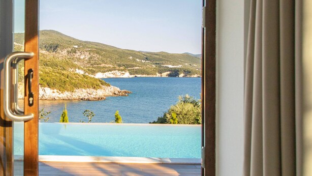 A view from a room with a wooden floor and a door, overlooking a pool and a body of water with hills in the background. - Zavia Villa Alpha 2B Syvota (Igoumenitsa)