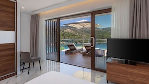 A modern hotel room with a large TV, a bed, a seating area with a table and chairs, and a balcony with a view of the landscape. - Zavia Villa Alpha 2B Syvota (Igoumenitsa)