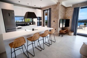 A modern kitchen with a large island, bar stools, a flat-screen TV, and a view of the sea. - Smirties Villa Haris 2bed (Lefkada)