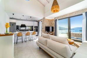 A modern living room with a sofa, a TV, and a stone fireplace. There is a kitchen in the background with bar stools. - Smirties Villa Haris 2bed (Lefkada)