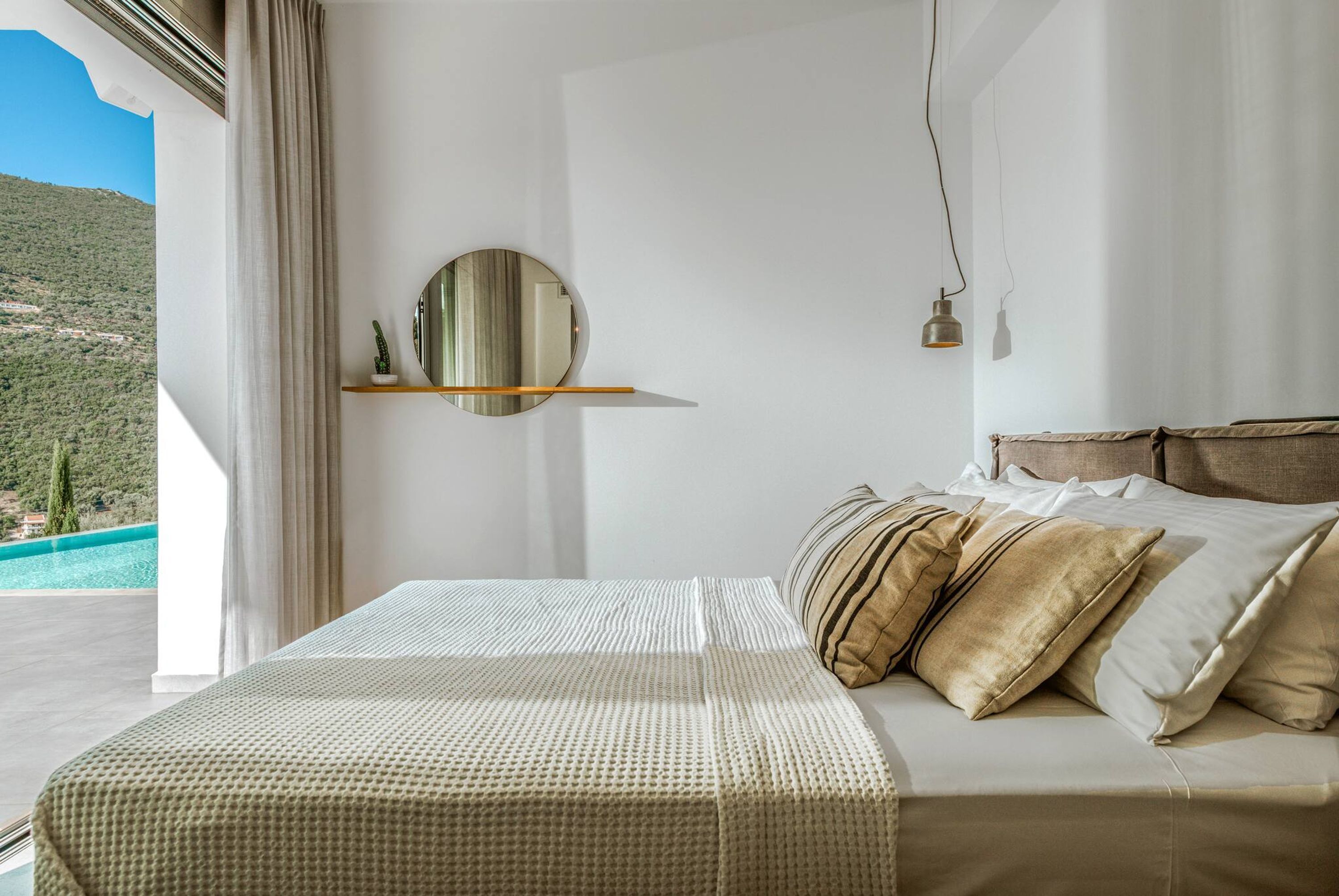 A bedroom with a bed, pillows, a round mirror, and a view of a mountainous landscape.