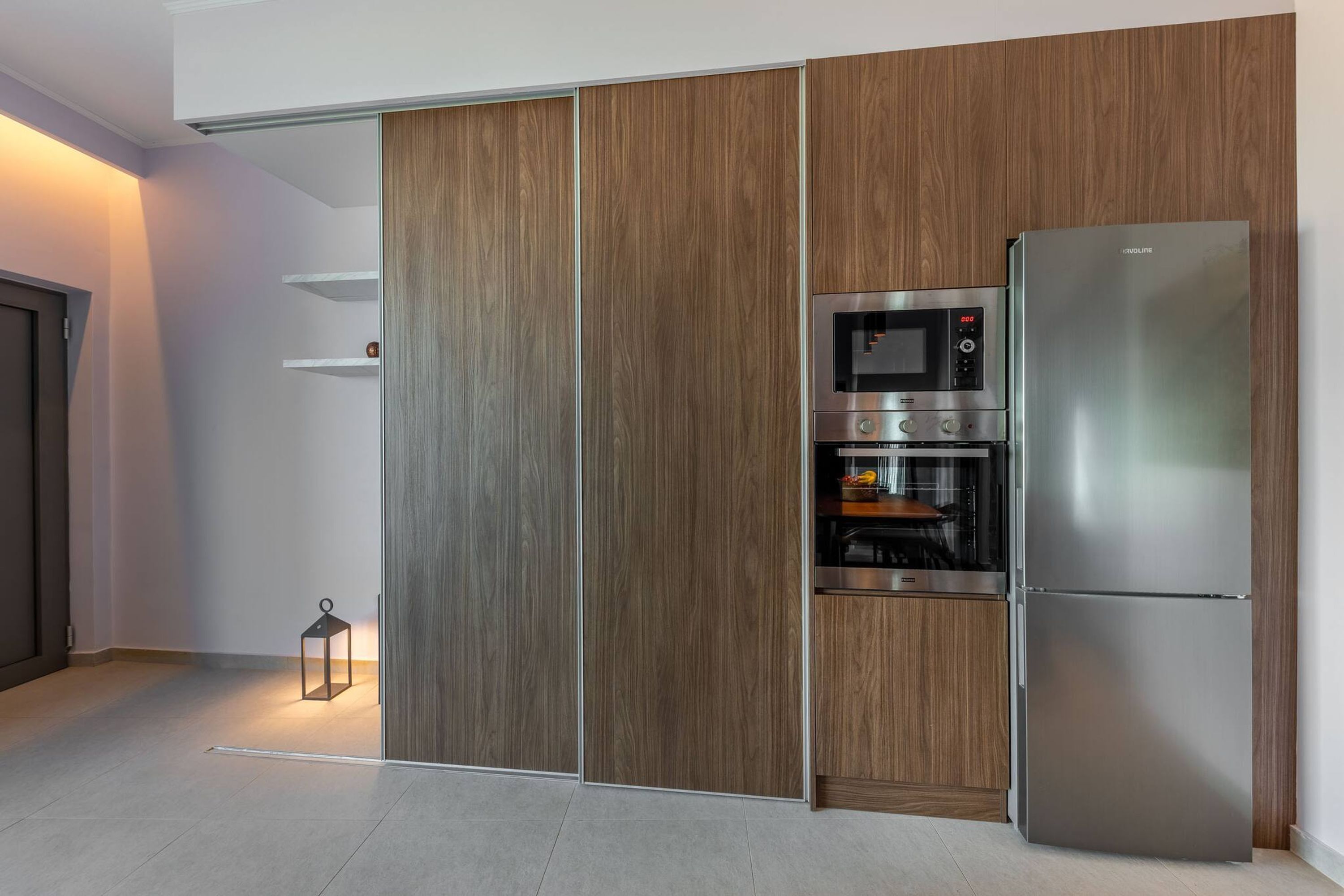 A modern kitchen with a built-in oven, microwave, and refrigerator, wooden paneling, and a small table with a lantern.