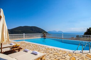 A swimming pool with a stone deck, lounge chairs, and a view of the sea and mountains. - Dream Villa in Ponti (Lefkada)