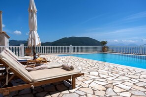 A poolside area with lounge chairs, a sun umbrella, and a view of mountains. - Dream Villa in Ponti (Lefkada)