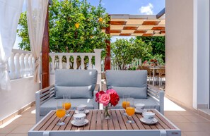 A sunlit outdoor seating area with two grey sofas, a wooden table with breakfast items, and a white fence with greenery behind. - Dream Villa in Ponti (Lefkada)