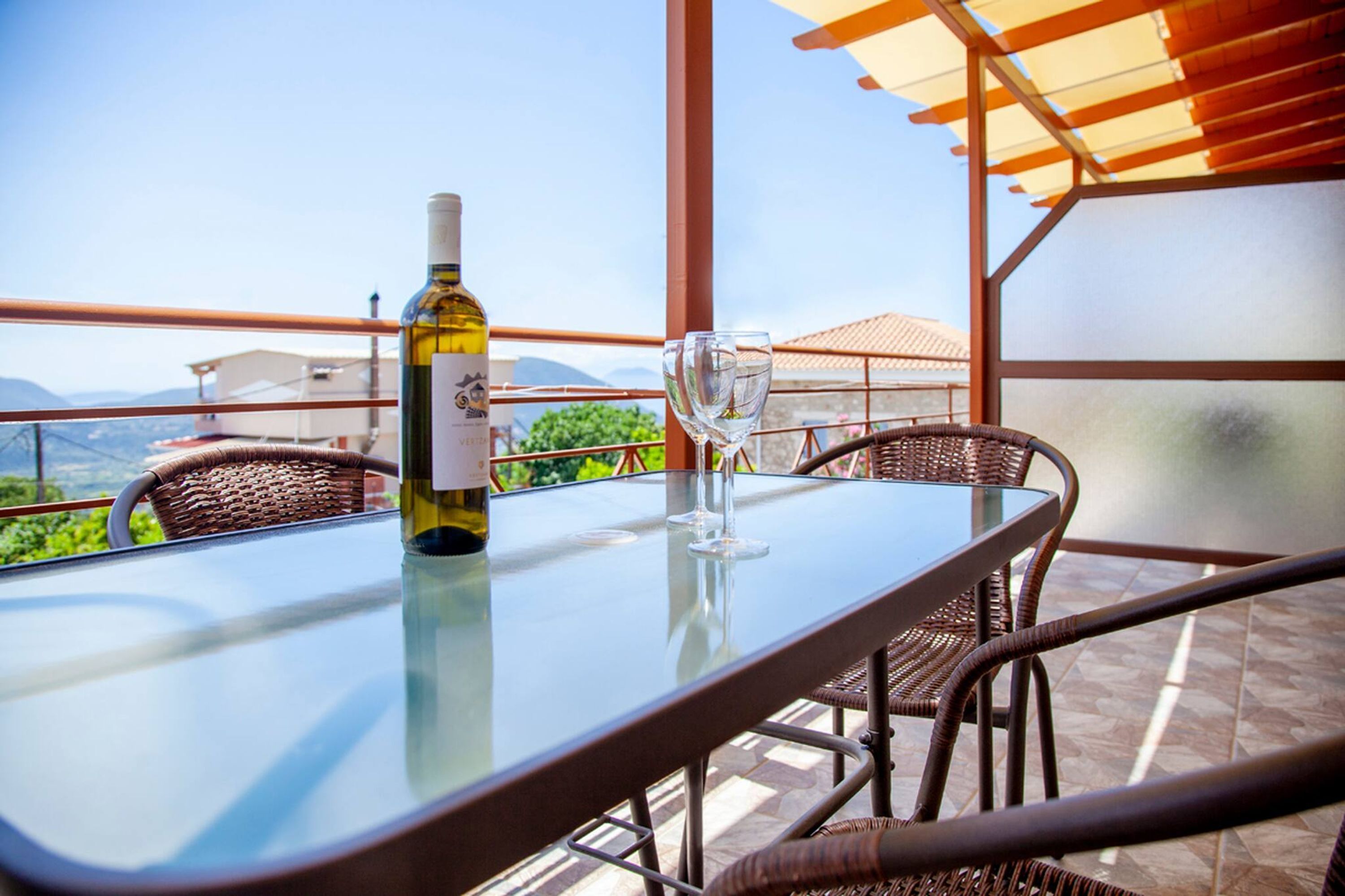 A glass table with a bottle and glasses, wicker chairs, and a view of the landscape.