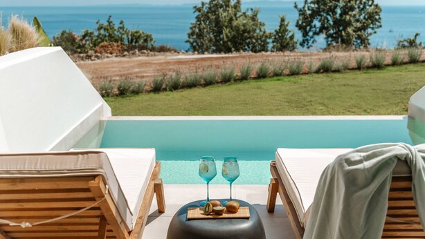 Blick auf einen Pool mit Liegestühlen, einem Tisch mit Getränken und den Meerblick.