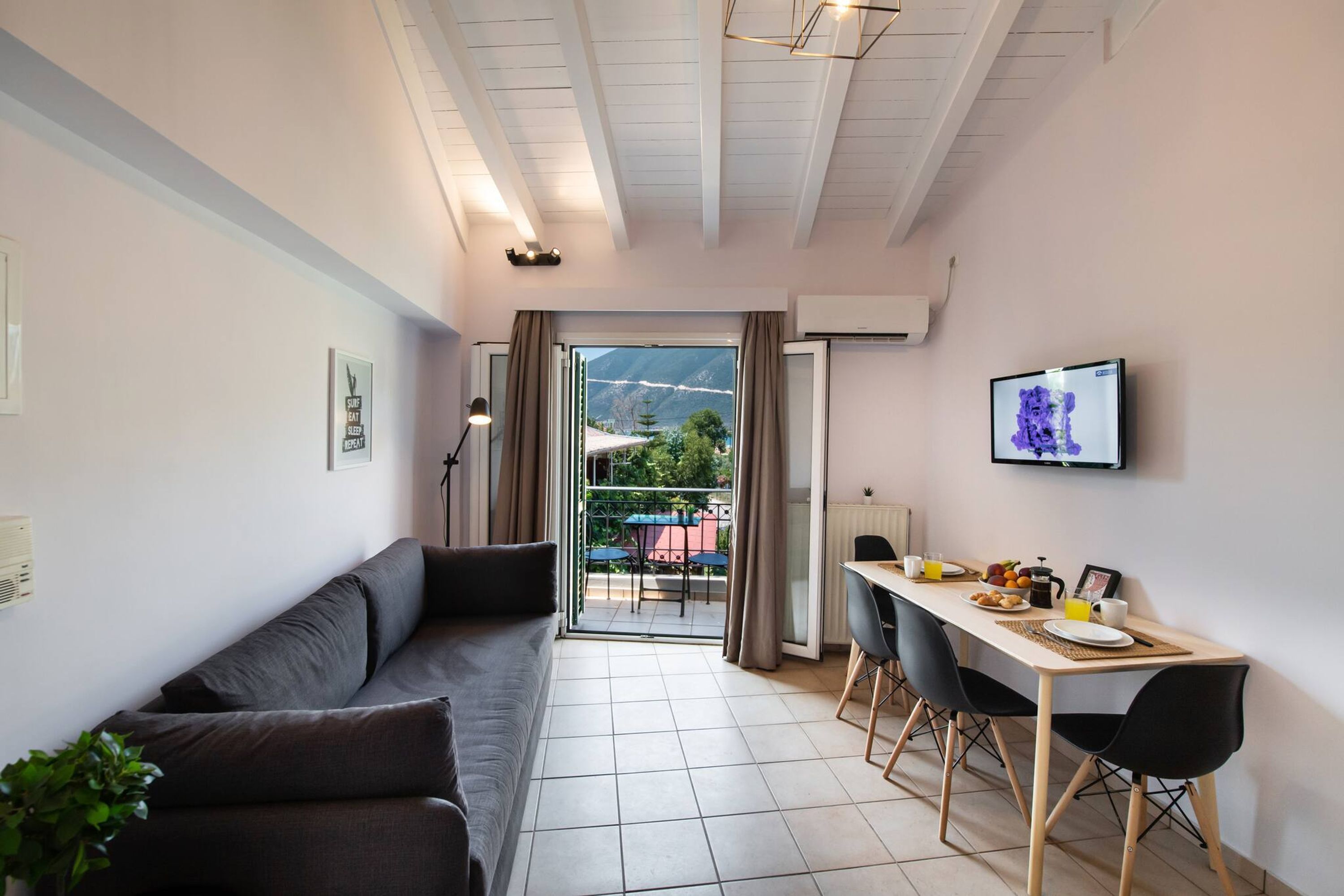 A modern living room with a sofa, a dining table with chairs, a TV, and a view of the outdoors.